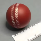 A Promotional Stress Ball Cricket Red is placed on a flat surface next to a white ruler with centimeter and millimeter markings. Its prominent stitching adds an elegant touch, making it ideal for promotional displays.