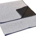 A folded picnic blanket with a geometric pattern in white and gray on one side and solid black on the other. It has a blue trim around the edges. The blanket is spread out on a white background.