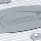 Close-up of a silver metal surface with an oval plaque featuring the text Petersen & Holmes. The metal has a ribbed texture and is fastened with bolts.
