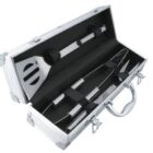 An open silver metal case containing barbecue tools on black padding. The set includes a spatula, tongs, and a fork, all with metallic handles. The case has a handle and latches for closing.