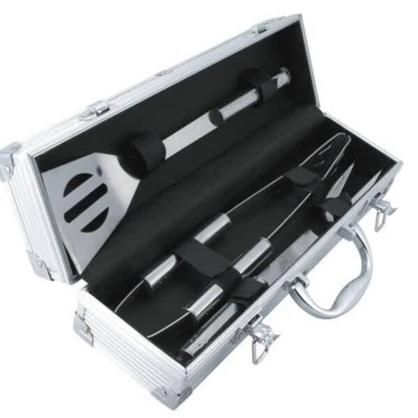 An open silver metal case containing barbecue tools on black padding. The set includes a spatula, tongs, and a fork, all with metallic handles. The case has a handle and latches for closing.