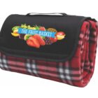 A folded red and black plaid blanket with a black flap featuring a logo for The Fruit Basket, decorated with images of various fruits. The blanket has a handle on top for easy carrying.