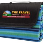A folded striped blanket with blue, green, and black horizontal lines. The black carrying flap shows THE TRAVEL CAMP logo with an image of a horse-drawn carriage and text GATEWAY TO FLAMING GORGE.