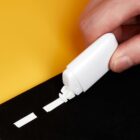Hand using correction tape on black paper against a yellow background.