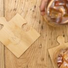 Two glasses of iced drinks are placed on a wooden table alongside puzzle-shaped wooden coasters. One hand holds a glass. The wood has a natural grain pattern, and the coasters are engraved with circular designs.