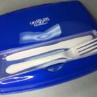 Blue lunchbox with clear lid section holding a white plastic fork and knife.