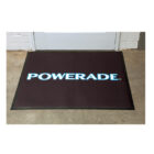 A rectangular black floor mat placed in front of a door, featuring the word POWERADE in bold white and blue letters. The mat is on a grey concrete floor against a white brick wall.