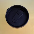 Close-up of a black travel mug lid with a closed drinking spout, set against a gradient yellow background.
