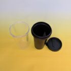Two cups are placed on a yellow surface. On the left is a transparent plastic cup, and on the right is a black travel mug with its lid sitting next to it. The background transitions from yellow to a lighter shade.