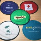 Five foldable frisbees with logos arranged on a wooden surface: JewishCare, Australian Red Cross, YoGo, EQI, and LiveLighter. Each frisbee is a different color and features its respective organizations branding.