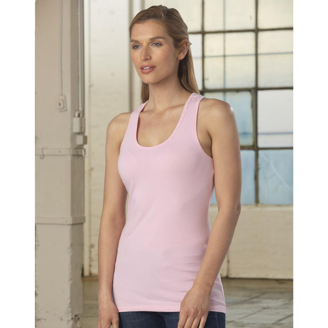 A woman wearing a Ladies Fitted Stretch Company Singlet T-shirt and jeans stands indoors by a frosted glass window. She gazes to the side, surrounded by exposed white beams and a light, neutral color scheme.