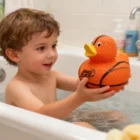 A smiling child enjoys bath time with a Basketball Duckies toy in hand.