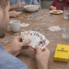A person holds the Deck Of Cards at a table with puzzles, food, and a yellow box.