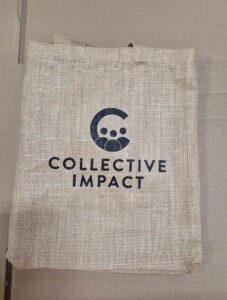 Natural jute tote bag printed with the Collective Impact logo in black.