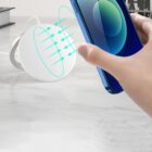 A hand holding a blue smartphone near a wireless charging pad on a marble surface. Green arrows between the phone and charger illustrate the wireless charging process. A stack of books is partially visible in the background.