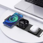 A wireless charging station on a white surface holds a smartphone displaying a charging icon, a smartwatch, and wireless earbuds. A laptop is partially visible in the background.