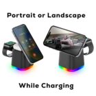 A multi-device charging stand with LED lights holds a smartphone in portrait mode on the left and landscape mode on the right. It also charges a smartwatch positioned on the side. Text reads, Portrait or Landscape While Charging.