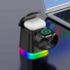 A sleek black charging station with a colorful LED base charges an AirPods case and a smartwatch. A laptop is partially visible beside it on a glossy dark surface.