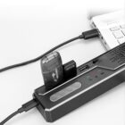 A vaping device inserted into an USB charging station connected to a laptop. The station has LED indicators, a power button, and volume controls, with cables plugged in. The laptop partially visible on the right.