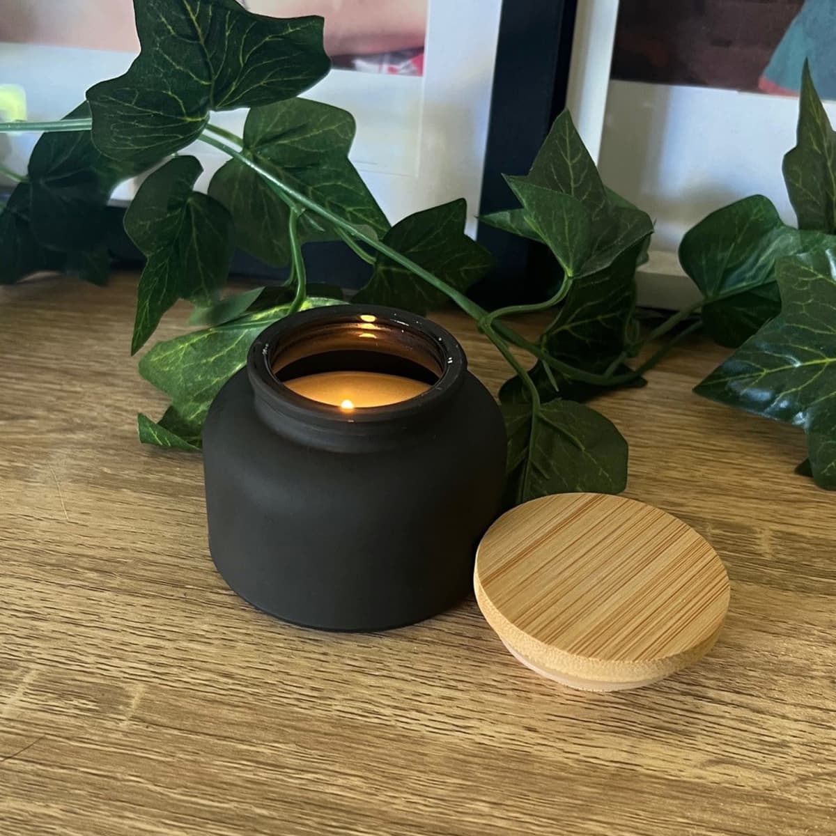A lit candle in a black jar with a wooden lid beside it, on a wooden surface with green leaves.