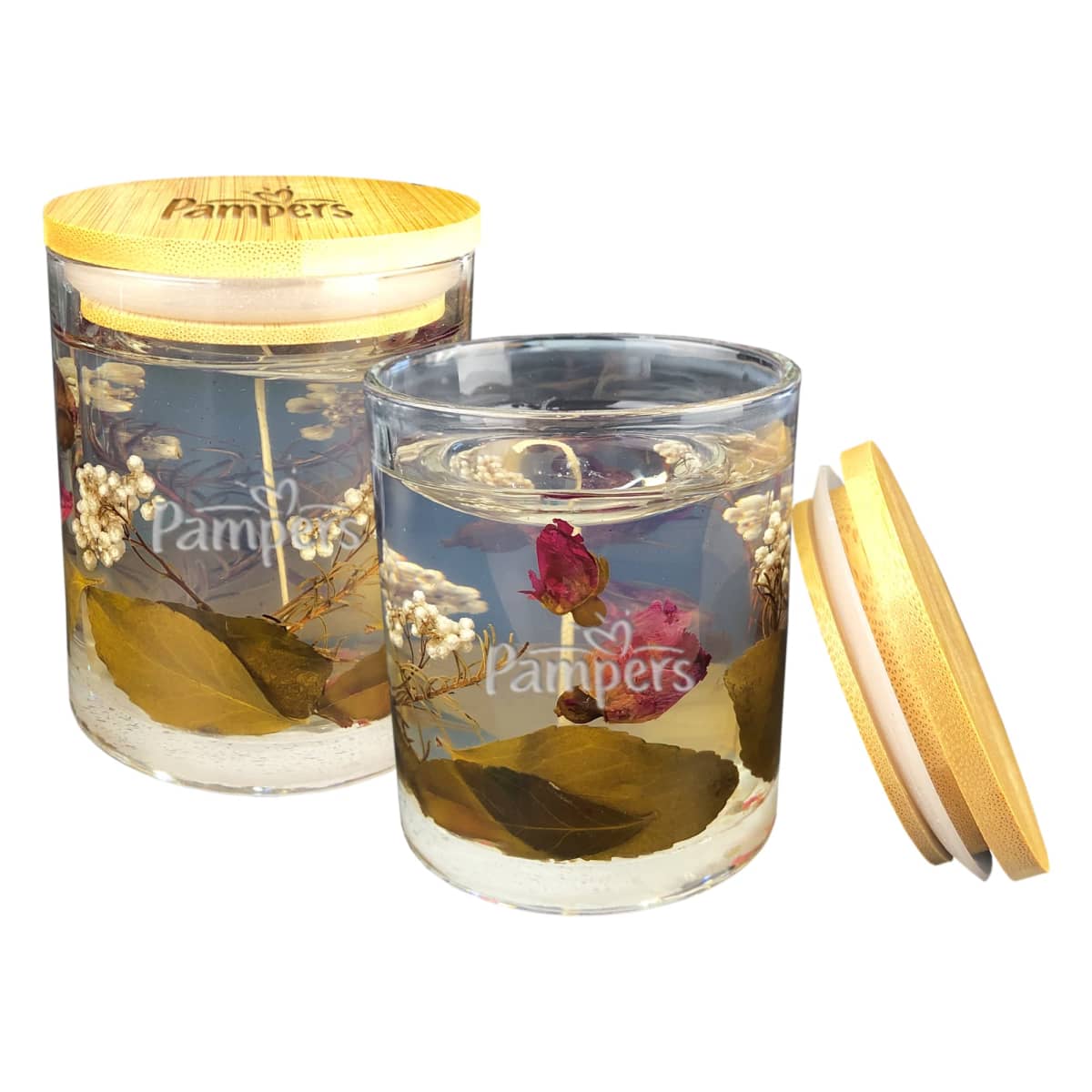 Two decorative candles in glass jars with wooden lids, featuring embedded dried flowers and leaves. The Pampers logo is visible on the front. One lid is placed beside its jar.