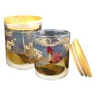 Two glass candles with wooden lids, featuring embedded dried flowers and leaves. One candle is open, while the other is closed. The arrangement includes pink, white, and green botanical elements in a clear wax base.