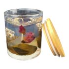 A transparent candle with embedded dried flowers and leaves, including a prominent pink rose, sits next to its light-colored wooden lid. The candle is made of clear gel, showcasing the foliage within.