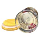 A glass jar candle lies on its side with a wooden lid removed beside it. The jar is filled with clear wax and embedded dried flowers, creating a decorative appearance.