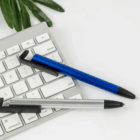 A Plastic Stylus Phone Stand and two pens sit on a white keyboard with a green leaf nearby.