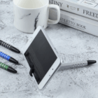 A smartphone on a Metal Stylus Phone Stand sits beside pens, books, and a marble mug.