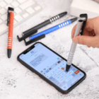 A hand uses the Metal Stylus Phone Stand by a keyboard and pens on a desk.