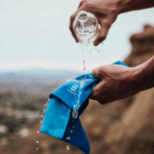 A hand pours water on a blue Everglades double layer cooling towel outdoors, blurred landscape behind.