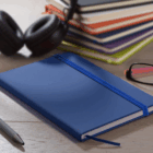 Rubber feel cover A5 notebook with pen, glasses, headphones; stacked notebooks behind on desk.