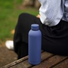 Blue water bottle on a wooden bench beside a person wearing black pants and a denim jacket.