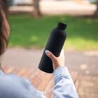 Person holding a black reusable water bottle outdoors near a park bench.