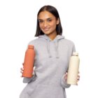 Woman in gray hoodie holding an orange bottle in one hand and a beige bottle in the other.