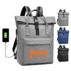 Gray backpack with a front pocket and a top flap secured by a black buckle. Has a USB charging port shown with a connected phone. Available in gray, blue, and black. HIKE for MENTAL HEALTH is printed in orange on the front pocket.