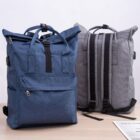 Two backpacks on a table: one blue with a visible front pocket and black straps, and one gray shown from the back with black straps. A magazine or brochure is partially visible on the table.
