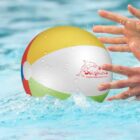 A colorful beach ball in a pool, splashed water droplets, hands reaching for it.