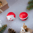 Two Christmas Stress Toy Santas Printed on a festive surface with pine cones and gift boxes.