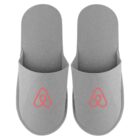 A pair of gray slippers with a red Airbnb logo on each slipper.