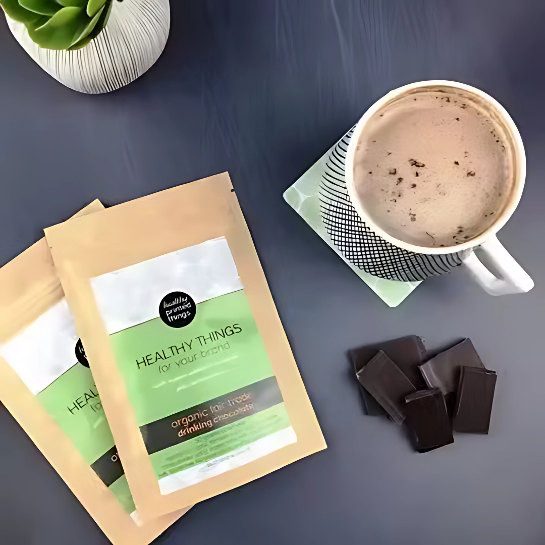 Two packets of Promotional Organic Drinking Chocolate, a mug, chocolates, and a plant on a gray table.