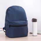A navy blue backpack and a white travel mug on a light wooden desk.