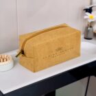 A gold-colored makeup bag with a zipper sits on a bathroom countertop. The bag is labeled Claire Austin Beauty with a small flower logo. In the background, theres a soap dish with white and beige soap beads and a faucet with a white flower.