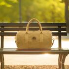 A tan duffel bag sits on a park bench in sunlight with a blurred green background.