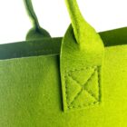 Close-up of a vibrant green felt tote bag. The focus is on the handle and stitching details, where the handle is attached to the main body of the bag with visible cross-stitching. The entire image is well-lit, highlighting the bags texture.