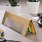 A cardboard triangular desk organizer holds a translucent ruler and colorful sticky notes in shades of blue, orange, and green. Its placed on a wooden surface near a potted plant and a pair of scissors.
