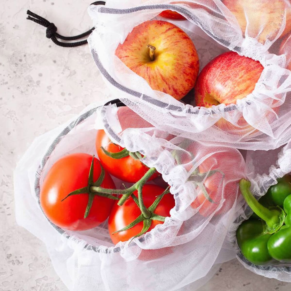Apples, tomatoes, and green bell peppers in reusable mesh produce bags on a light surface.
