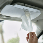 A hand pulls a tissue from a Super Sun Visor Logo Tissue Holder attached to a car visor.
