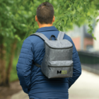 A person wearing a blue quilted jacket walks along a path lined with trees. They have a gray backpack with a small logo and the text MOUNTAIN SEEKERS on it. The background is blurred, focusing on the backpack and the person.